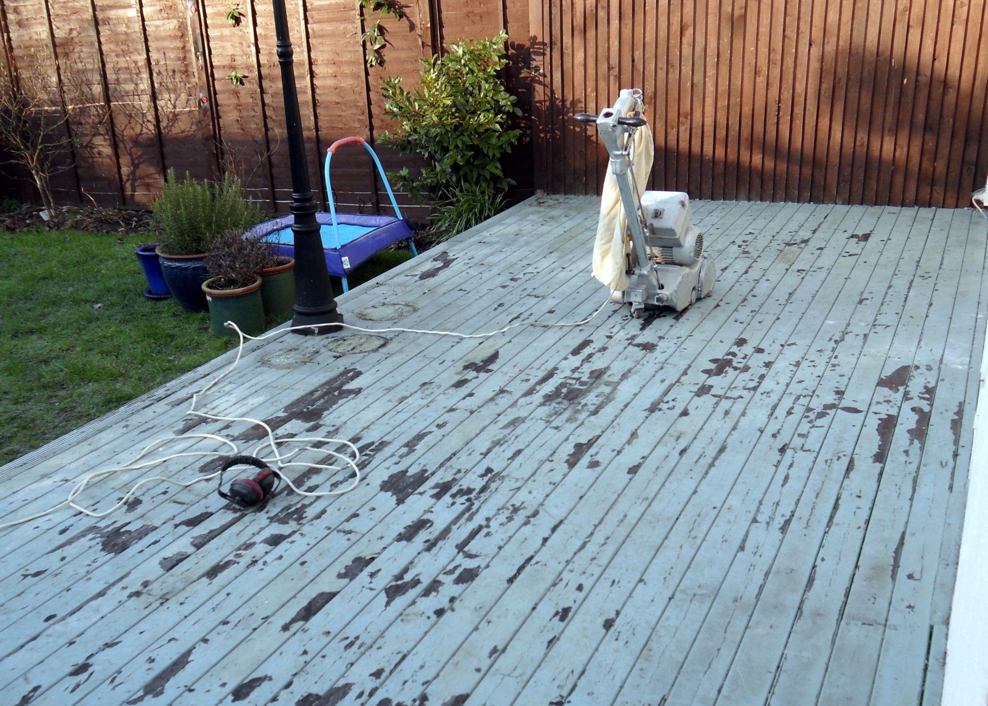Painted softwood timber decking floor in a Chelsea garden prepared for Deck Sanding, with a floor sander and safety headphones on the worn, peeling boards.